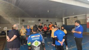 Uma imagem de um grupo de pessoas em uma quadra, participando de um treinamento esportivo. A composição da cena mostra um grupo de atletas, posicionados em um círculo, com um professor no centro e um pouco mais para trás do grupo. Aparentemente, todos estão em uma posição de atenção ou escuta.  O ângulo da imagem é de cima para baixo, o que ajuda a capturar toda a cena. Há uma presença clara de organização e concentração.