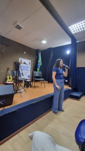 Uma mulher fala em um palco durante um evento, segurando um microfone, em um ambiente interno com decoração sóbria e uma bandeira brasileira. a mulher tem cabelos castanhos, usa uma blusa azul e calças de tecido solto da mesma cor. Ela segura um microfone, aparentemente falando ou cantando. Sua expressão facial parece neutra, concentrada na performance. A postura é elegante e relaxada, com os braços naturalmente posicionados. A vestimenta sugere um evento informal e a iluminação acentua seus traços e a roupa.