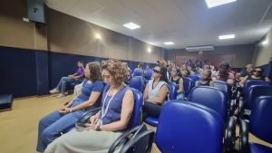 A composição da cena revela um auditório com fileiras de assentos azuis, preenchidas por diversas pessoas. A ação principal parece ser uma palestra ou apresentação, com as pessoas olhando para um ponto fora do enquadramento. A iluminação é artificial, com luzes no teto que iluminam o ambiente de forma uniforme. Os elementos estão posicionados de forma a dar uma ideia de profundidade, com as fileiras de cadeiras convergindo em direção ao fundo.

O público é diversificado, com pessoas de diferentes idades e gêneros. Alguns estão vestidos casualmente, enquanto outros parecem mais formais. As expressões faciais variam, com algumas pessoas parecendo atentas e outras mais relaxadas. Há algumas pessoas que estão usando óculos escuros, e outras que estão com seus olhos fechados.