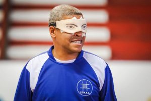 Sandro tem cabelo curto e grisalho, veste uma camiseta esportiva azul com detalhes brancos e o logotipo do ISMAC no peito. Ele está usando uma venda bege com olhos desenhados e fita adesiva branca presa ao rosto e à venda. A venda tem um elástico preto ao redor da cabeça para mantê-la fixa. Ele está em um ambiente interno, provavelmente um ginásio esportivo, com fundo desfocado de barras horizontais. Sandro aparece com expressão tranquila, boca parcialmente aberta, como se estivesse conversando ou explicando algo.